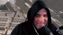 a man in a hooded sweatshirt is talking into a microphone with a sign that says ground in the background