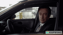 a man in a suit and tie is sitting in a car with the word kapwing on the bottom