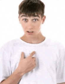 a young man wearing a white t-shirt is pointing at his chest .