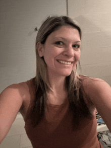 a woman in a brown tank top smiles while taking a selfie in a bathroom