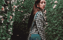 a woman wearing a plaid jacket and a blue bag stands in front of a wall of flowers