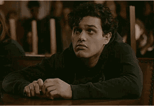 a young man is sitting at a table in a dark room with a group of people behind him .
