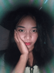 a girl with long black hair holds her hand to her face and looks at the camera