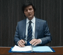 a man in a suit and tie is sitting at a desk looking at a folder .