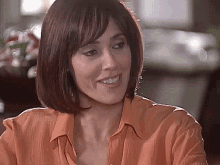 a woman with short brown hair is smiling and wearing an orange shirt .