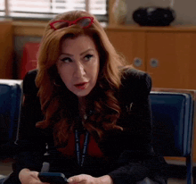 a woman with red hair and red glasses is sitting in a chair looking at her phone .