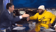a man in a yellow lakers sweatshirt is shaking hands with another man in a white hat .