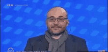 a bald man wearing glasses and a scarf is standing in front of a blue screen .