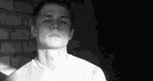 a black and white photo of a young man standing in front of a brick wall .