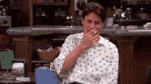 a man in a polka dot shirt is sitting in front of a counter covering his mouth with his hand .