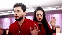 a man in a red jacket and a woman in glasses are standing next to each other on an airplane