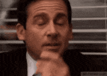 michael scott is sitting in front of a window with his eyes closed and his hand on his chin .