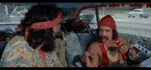 two men are sitting in a car and one has a bandana on his head that says ' hippie ' on it