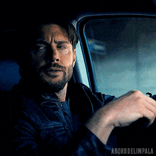 a man in a leather jacket is sitting in the driver 's seat of a car looking out the window