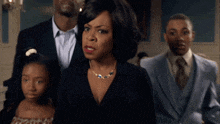 a woman with a necklace stands in front of a group of people in suits