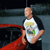 a bald man wearing a shirt that says colorado 's is standing next to a red car