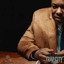 a man in a suit sits at a table with a trap city poster behind him