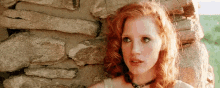 a woman with red hair and green eyes is leaning against a stone wall .