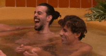 two men are laughing in a bathtub and one has a bun on his hair