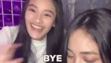 two young women are standing next to each other and smiling . one of the girls is saying `` bye '' .