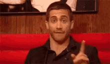 a man is sitting on a red couch making a funny face and pointing at something .