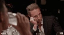 a man in a suit sits at a table with his head resting on his hand and # mafs written on the bottom right