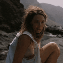 a woman in a white tank top sits on a beach with mountains in the background