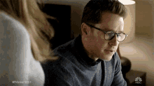 a man wearing glasses is talking to a woman with the nbc logo behind him