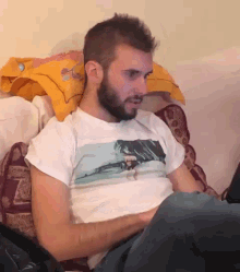 a man with a beard is sitting on a couch wearing a white t-shirt