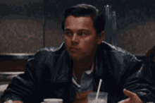 a man in a leather jacket is sitting at a table talking to another man .