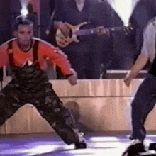 a man in overalls is dancing on a stage with a guitar player in the background .
