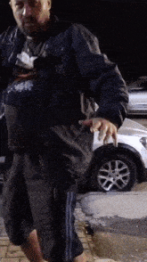 a man in a black jacket and grey shorts stands in front of a silver car