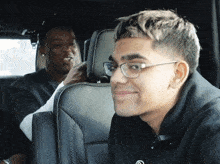 a man wearing glasses sits in the back seat of a car next to another man