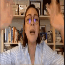 a woman wearing glasses is sitting in front of a bookshelf with her hands in the air .