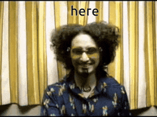 a man with curly hair wearing sunglasses and a blue shirt is smiling and says here
