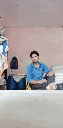 a man in a blue shirt sits on a bed with his feet crossed