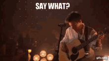 a man is playing an acoustic guitar and singing into a microphone while saying `` say what '' .