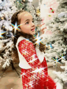 a little girl in a red dress and white vest stands in front of a christmas tree