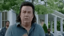 a man with long hair is standing in front of a white house making a funny face .
