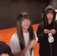 two girls are standing next to each other on a wooden floor . one of the girls is wearing a hoodie .