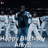 a man in a blue jacket stands in front of a group of dancers and says " happy birthday amy !! "