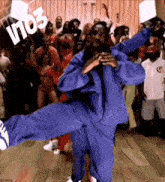 a man in a blue jumpsuit is dancing in front of a crowd with the letters v103 visible