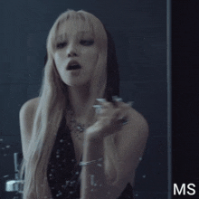 a woman with long blonde hair looks at herself in a mirror with the letters ms visible
