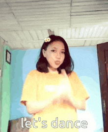 a girl in a yellow shirt is standing in a room and says let 's dance .