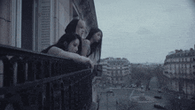 three women lean on a balcony railing overlooking a city street