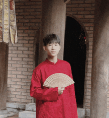 a young man in a red shirt holds a fan in front of a brick wall