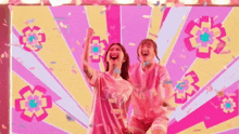 two women are standing next to each other in front of a pink and yellow background with confetti falling around them .