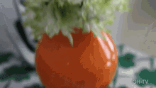 a close up of an orange vase with green flowers on a table