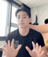 a young man wearing a black t-shirt is sitting in front of a window with his hands outstretched .