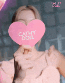 a woman is holding a pink heart that says cathy doll in front of her face